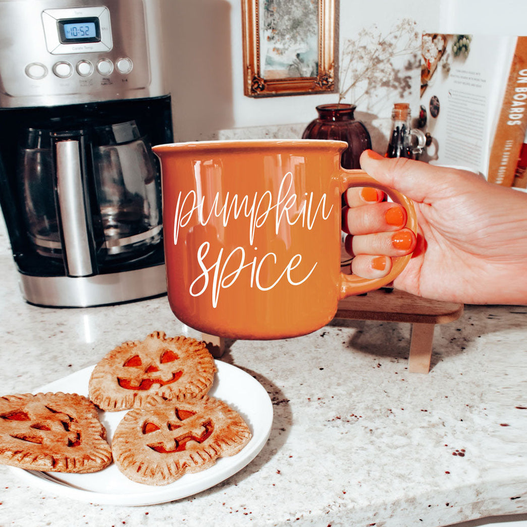 Person holding an orange 'Pumpkin Spice' mug with pumpkin pies on a kitchen counter.