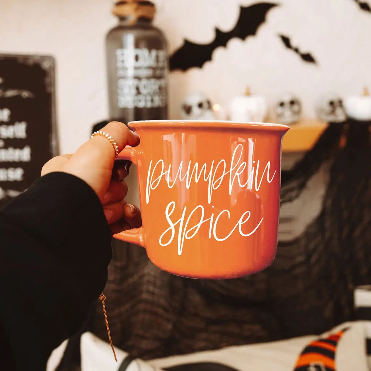 Person holding an orange 'Pumpkin Spice' mug with Halloween decorations in the background