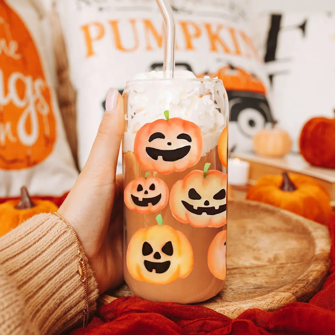 Glass with Halloween-themed pumpkin designs held by a person, surrounded by pumpkins and autumn decor.