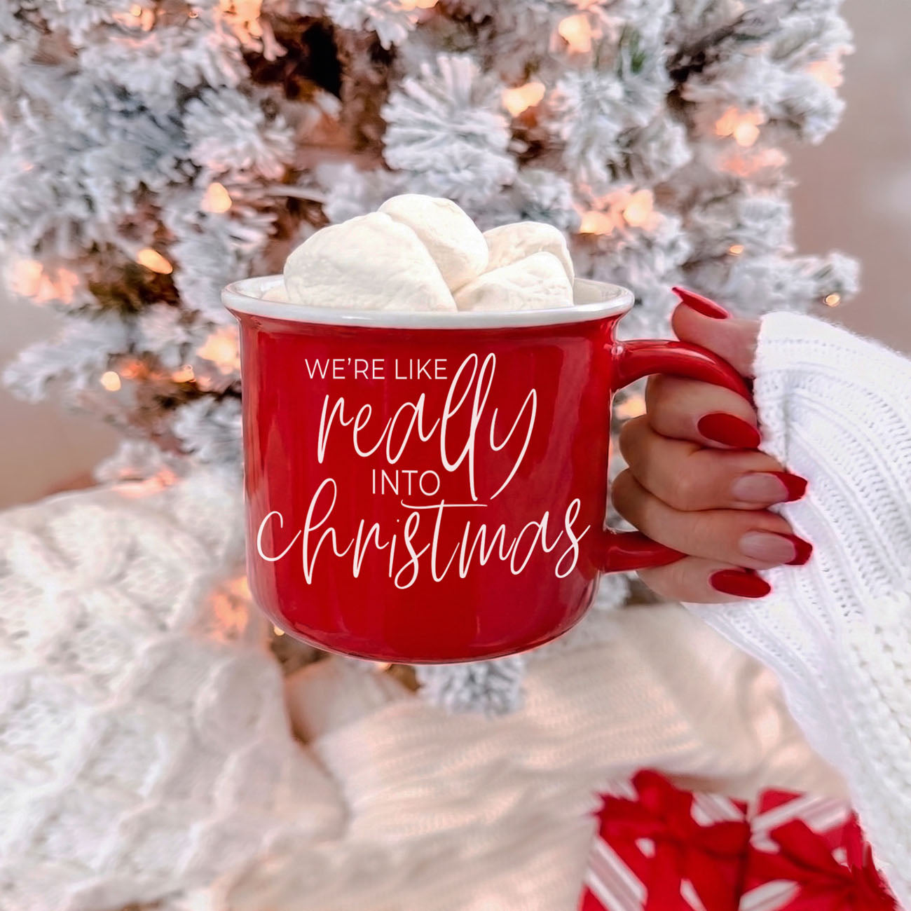 Red mug with 'We're like really into Christmas' text held by a person in front of a decorated Christmas tree.