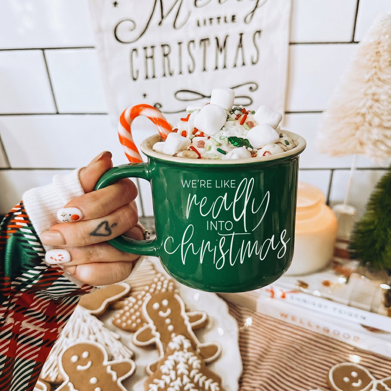 Green mug with Christmas-themed text held by a person, surrounded by festive decor.