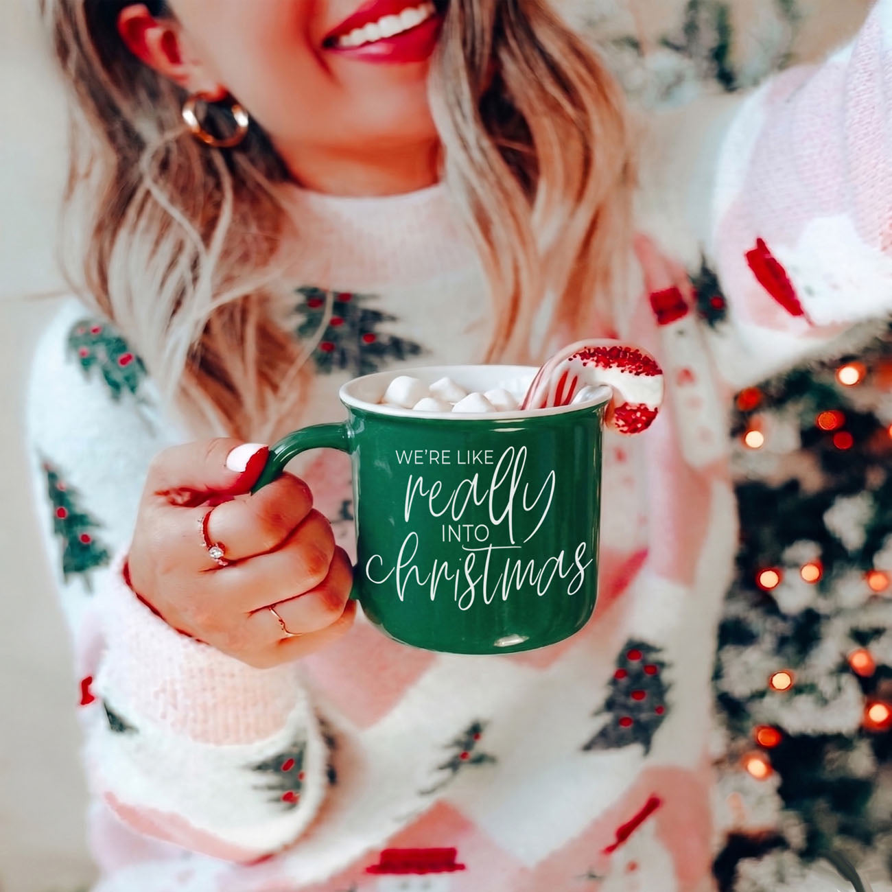 Person wearing a festive sweater holding a green mug with Christmas text, Christmas tree in the background