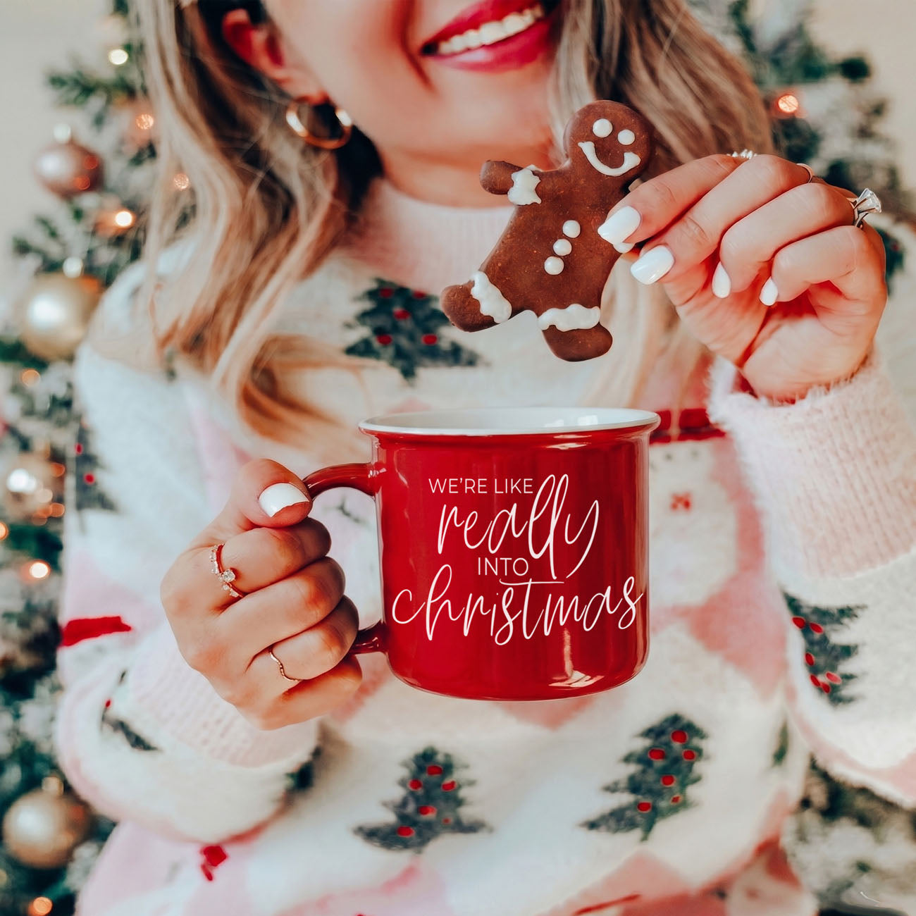Person holding a red mug with Christmas text and a gingerbread cookie in front of a decorated tree.
