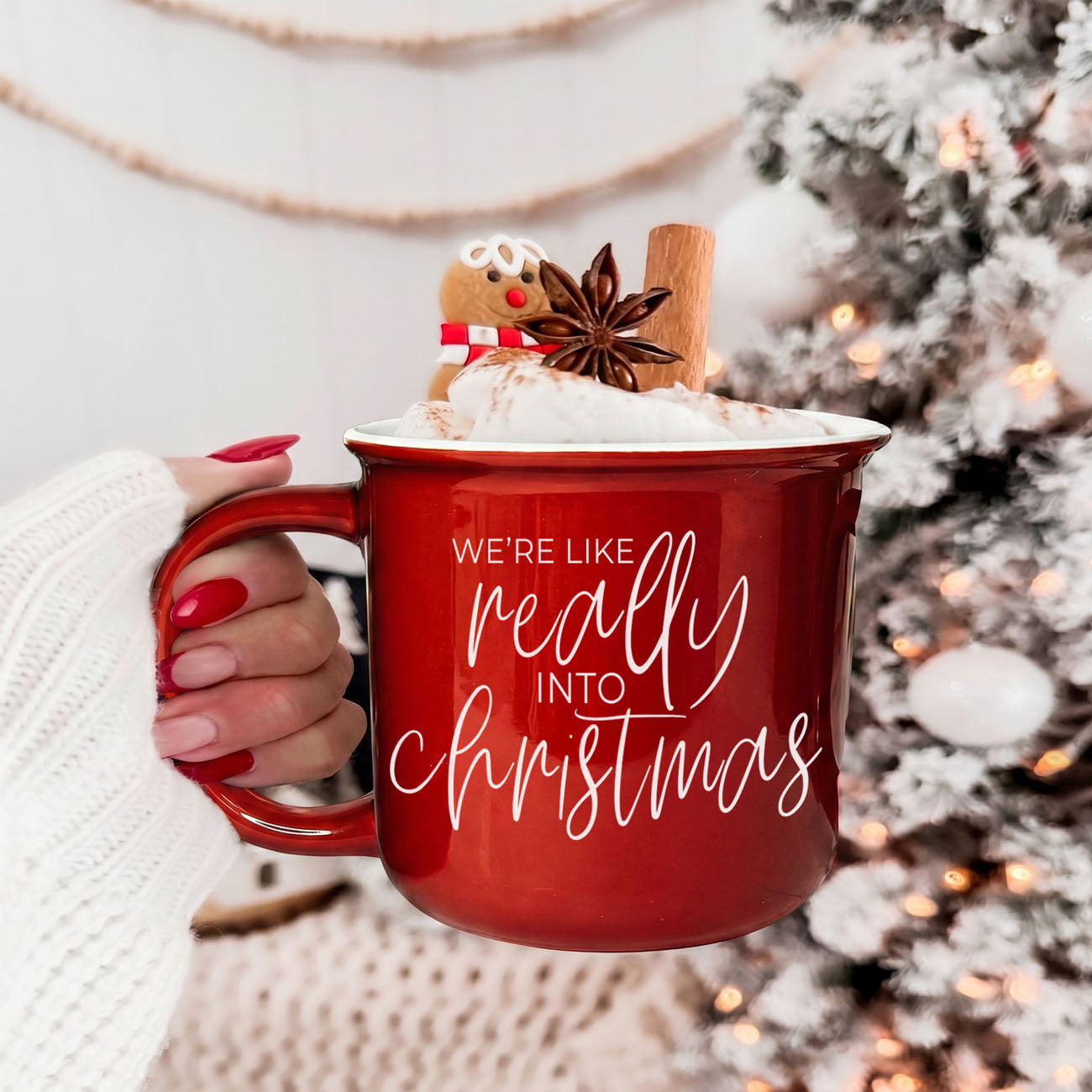 Red mug with 'We're like really into Christmas' text, held by a person, against a festive background with a Christmas tree.