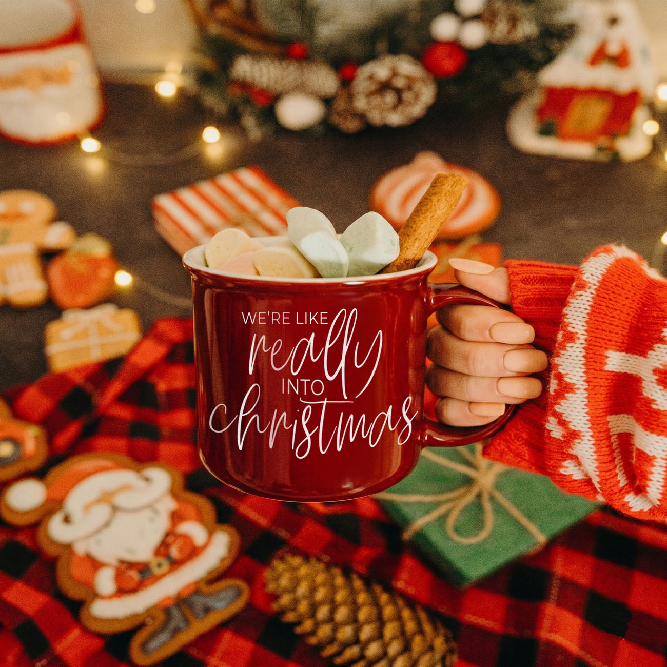 Red mug with 'We're like really into Christmas' text, held by a person in a red sweater, surrounded by Christmas decorations.