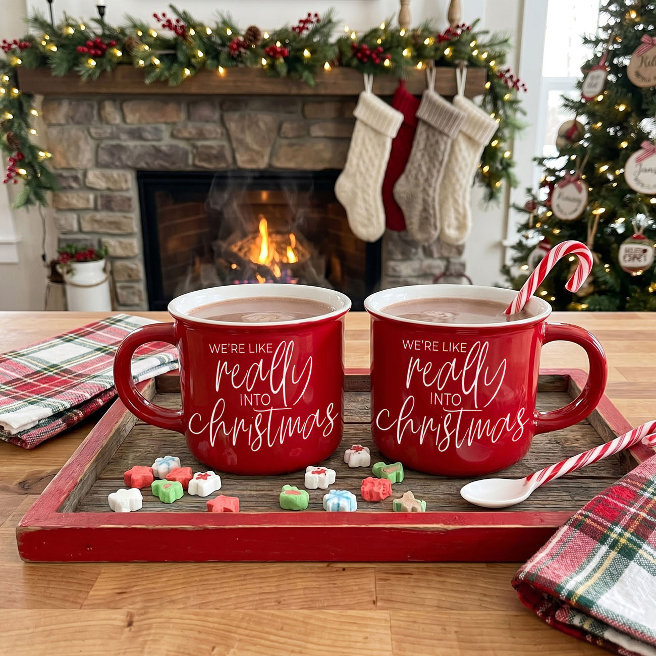 Two red mugs with Christmas-themed text on a wooden tray in front of a fireplace and Christmas tree.