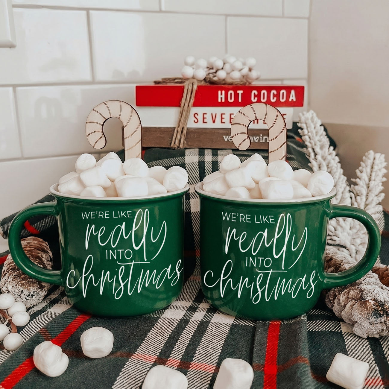 Two green mugs with Christmas text on a plaid blanket with marshmallows.