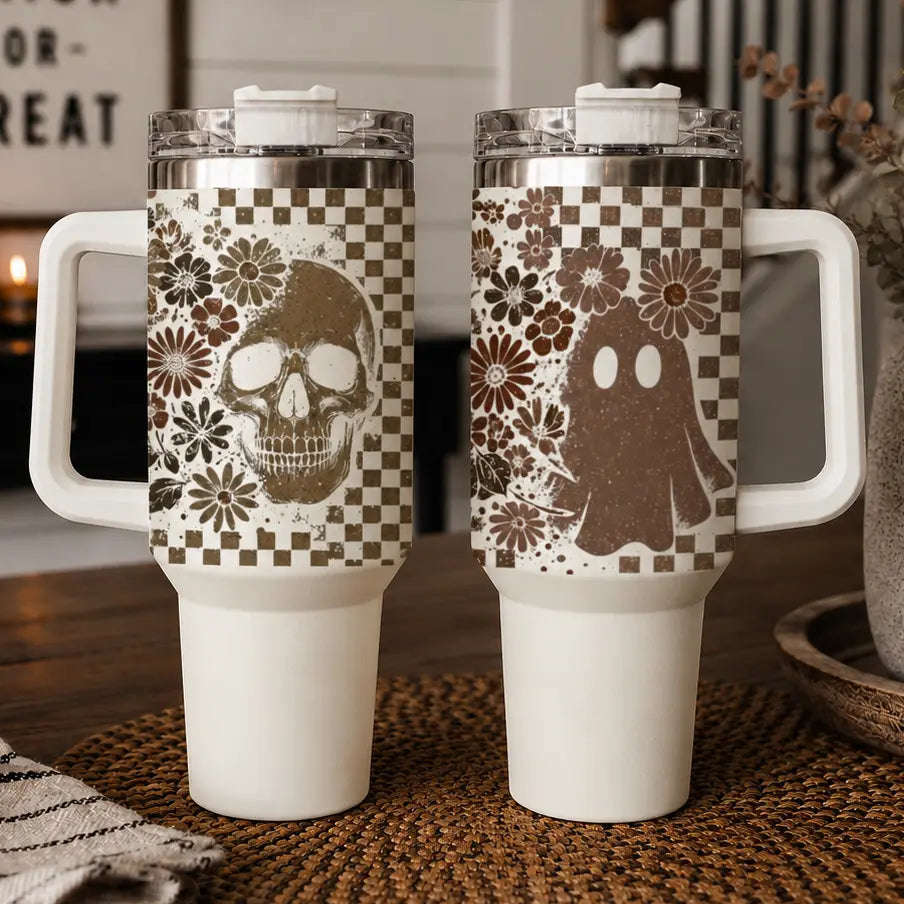 Two handled travel tumblers with clear lids and white bases, shown side by side on a woven brown mat. The left tumbler has a brown skull and floral print over a beige-and-white checkerboard pattern.