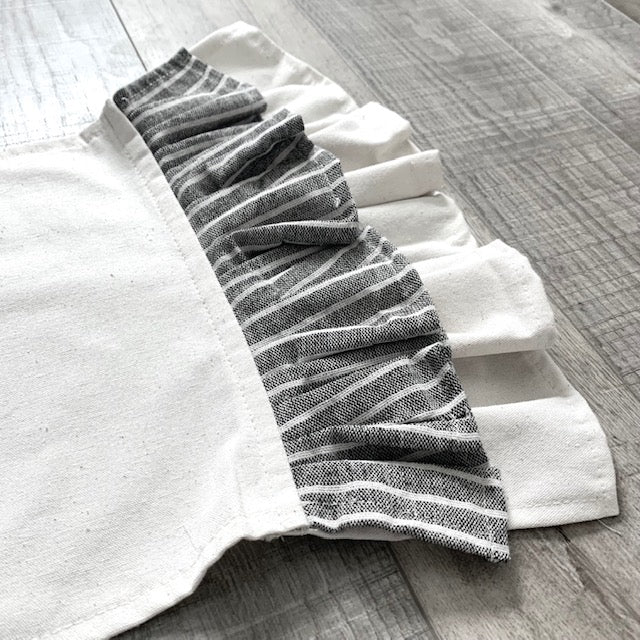 A white fabric apron or garment laid flat on a gray wood floor, featuring a ruffled trim made of gray and white striped fabric.