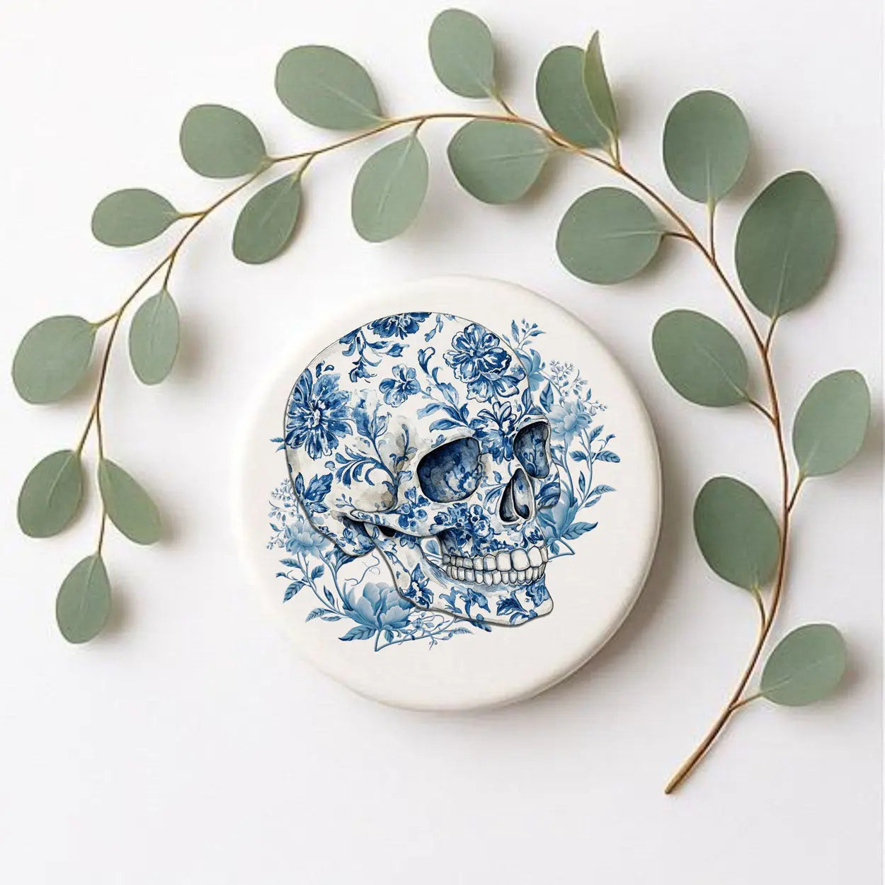 Decorative plate with blue floral skull design surrounded by green leaves on a white background