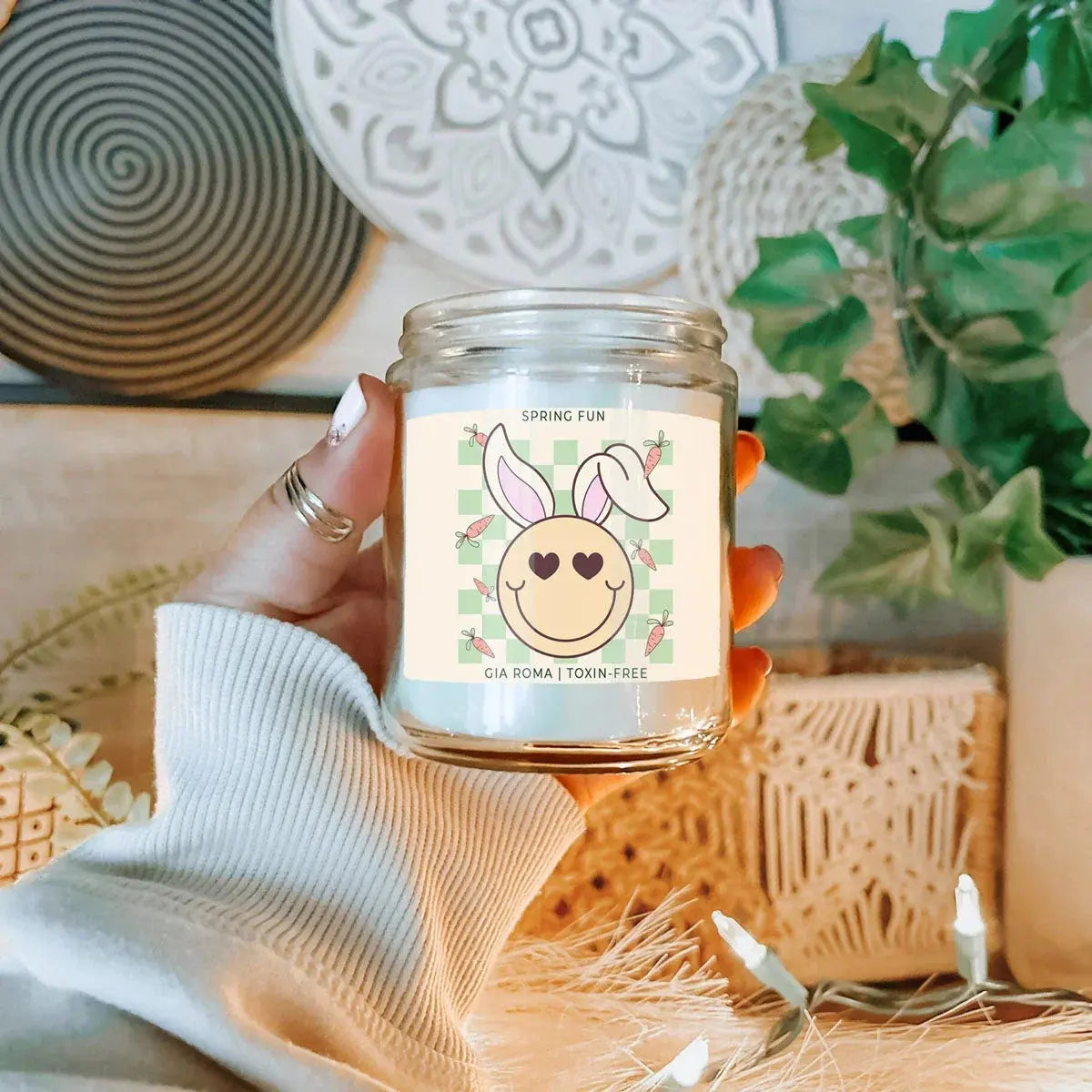 Candle jar with a decorative label held by a hand, with a blurred indoor background.