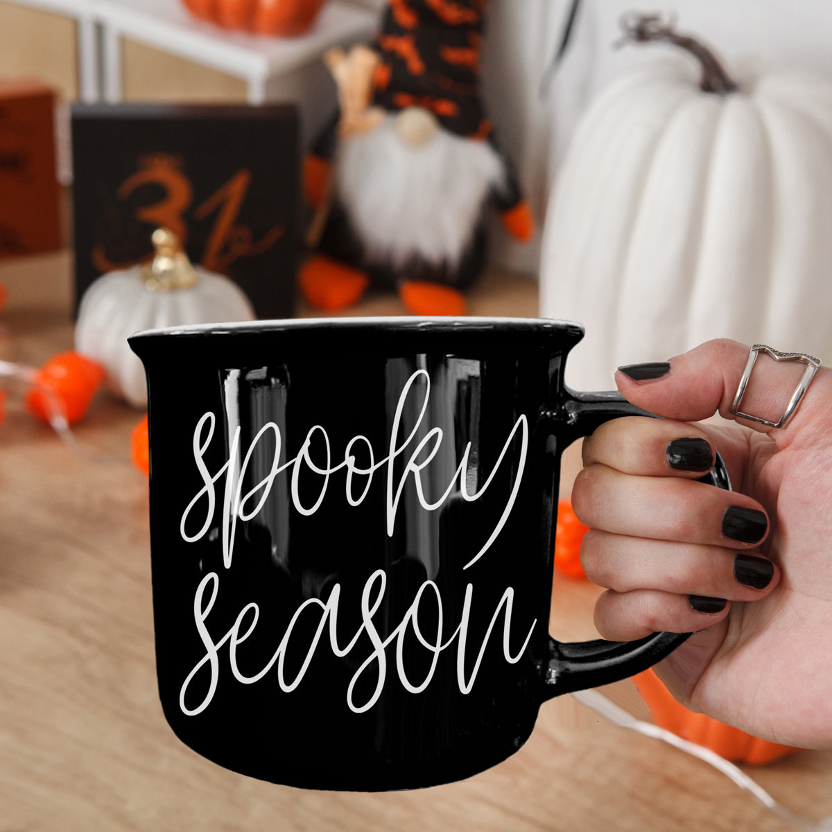 Black mug with 'Spooky Season' text held by a hand, with Halloween decorations in the background.