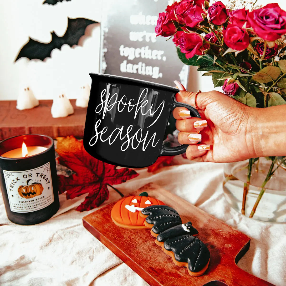 Hand holding a 'Spooky Season' mug with Halloween-themed decor including a candle, cookies, and flowers.