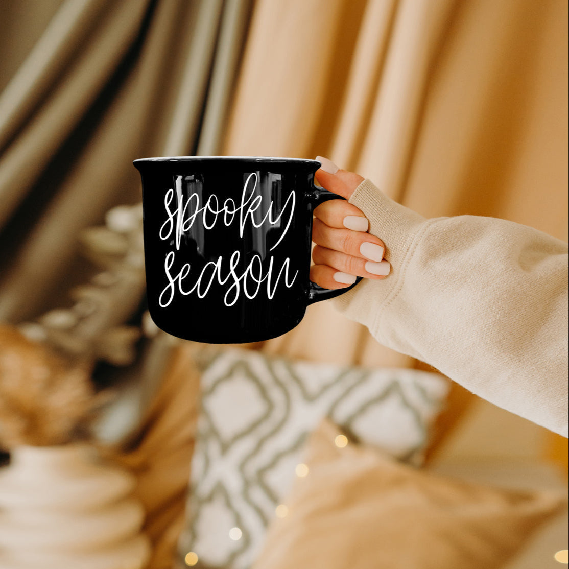 Black mug with 'Spooky Season' text held by a person in a cozy indoor setting.