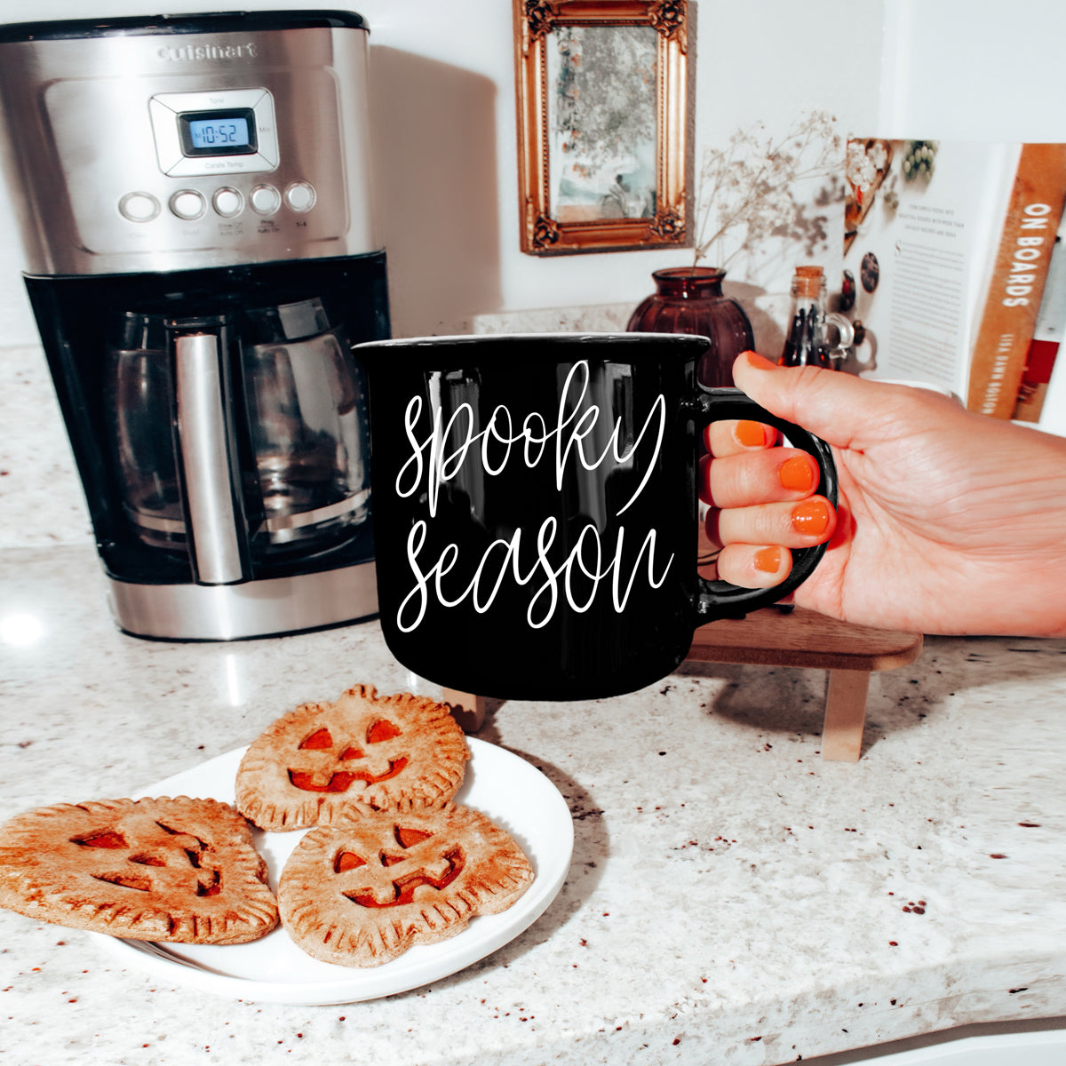 Person holding a 'Spooky Season' mug with pumpkin pies and a coffee maker in the background.