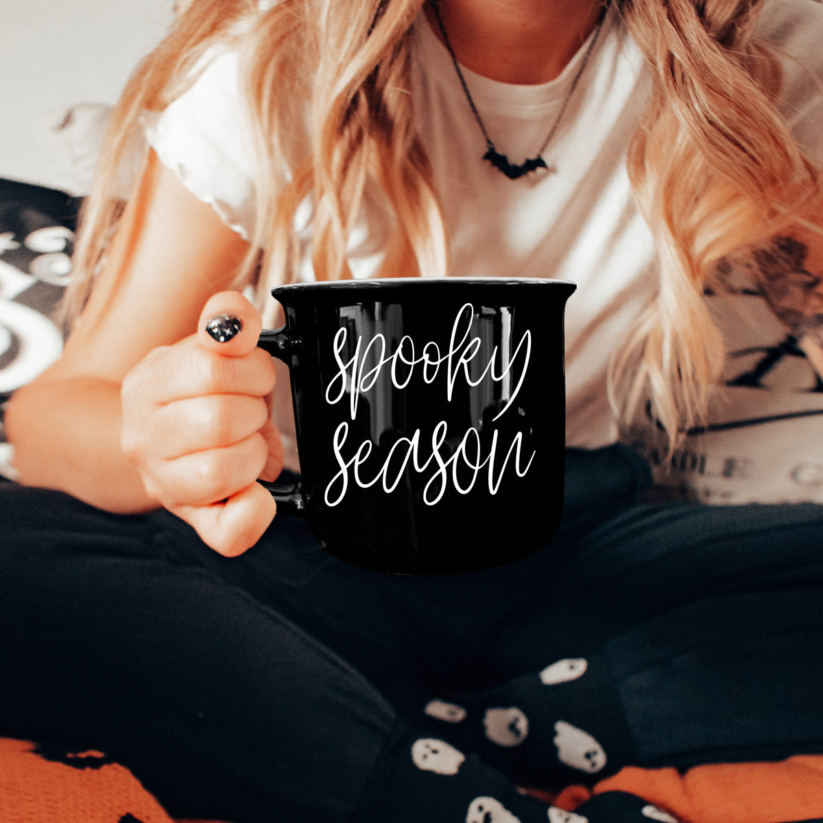 Person holding a black mug with 'Spooky Season' text, sitting on a Halloween-themed blanket.