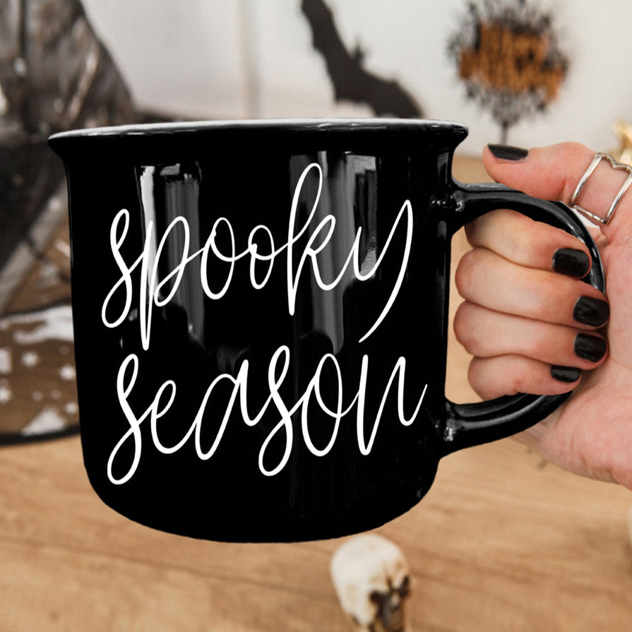 Black mug with 'Spooky Season' text held by a hand, Halloween-themed background