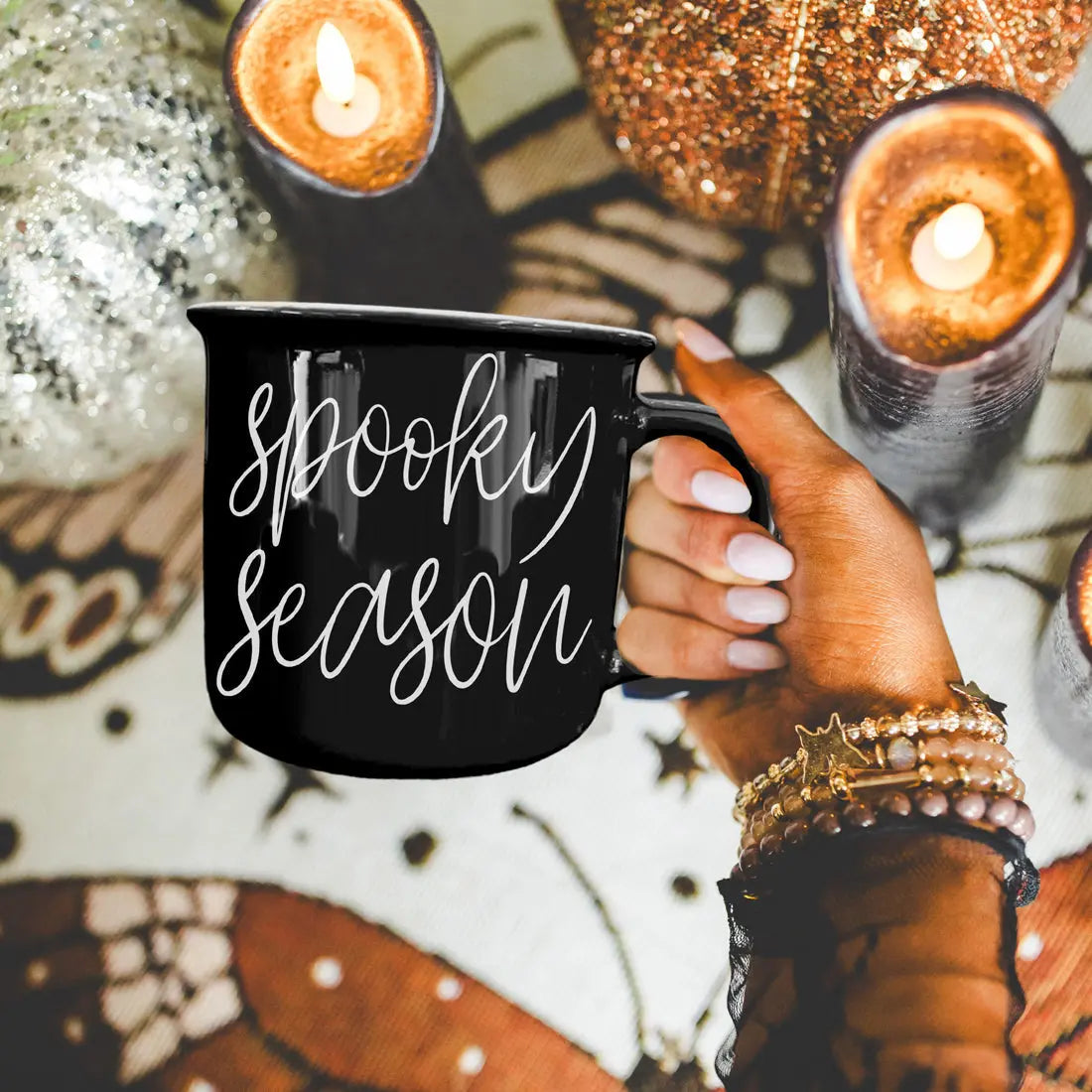 Black mug with 'spooky season' text held by a hand against a Halloween-themed background with candles and decorations.