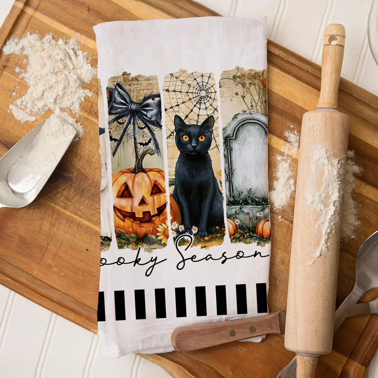 Halloween-themed kitchen towel with black cat, pumpkin, and tombstone design on a wooden surface with kitchen items.