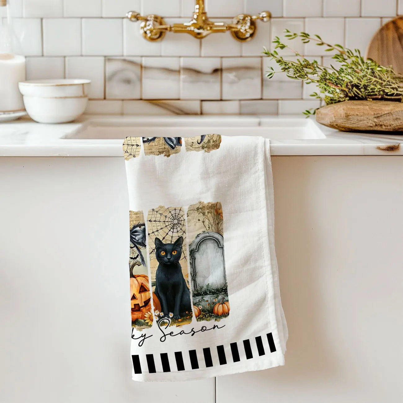 Decorative kitchen towel with Halloween design featuring a black cat, tombstones, and pumpkins hanging on a kitchen towel rack.