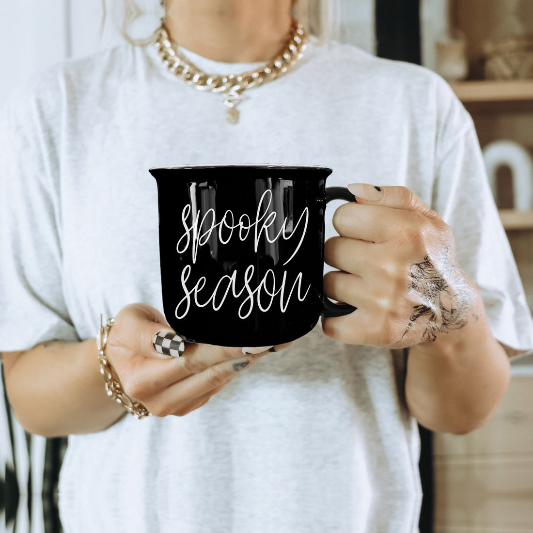 Person holding a black mug with 'Spooky Season' text, wearing a white shirt and gold jewelry.