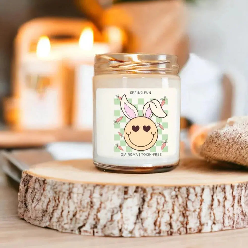 Candle in a glass jar with a decorative label on a wooden surface, with blurred candles in the background.