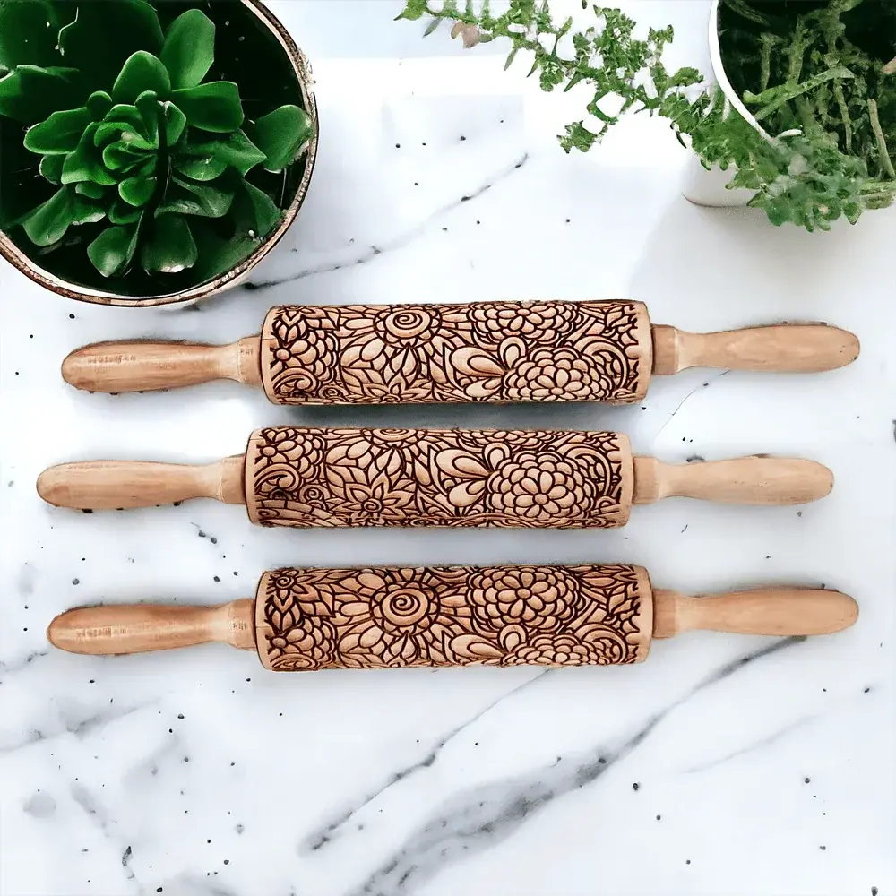Three wooden rolling pins with floral patterns on a marble surface with plants in the background.