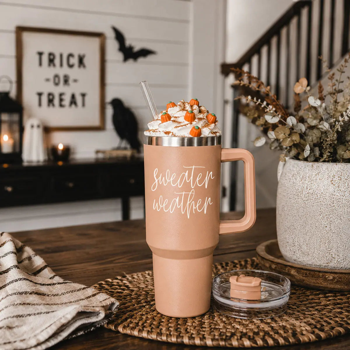 Pink travel mug with 'Sweater Weather' text, topped with whipped cream and candy pumpkins, on a wooden table.