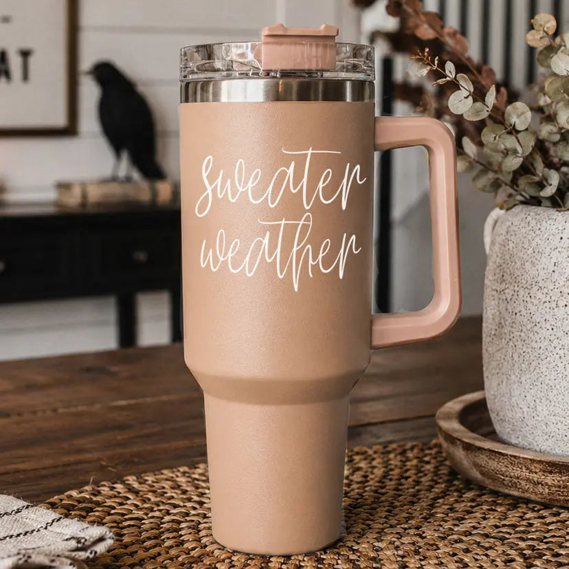 Beige travel mug with 'sweater weather' text on a wooden surface with decorative elements.