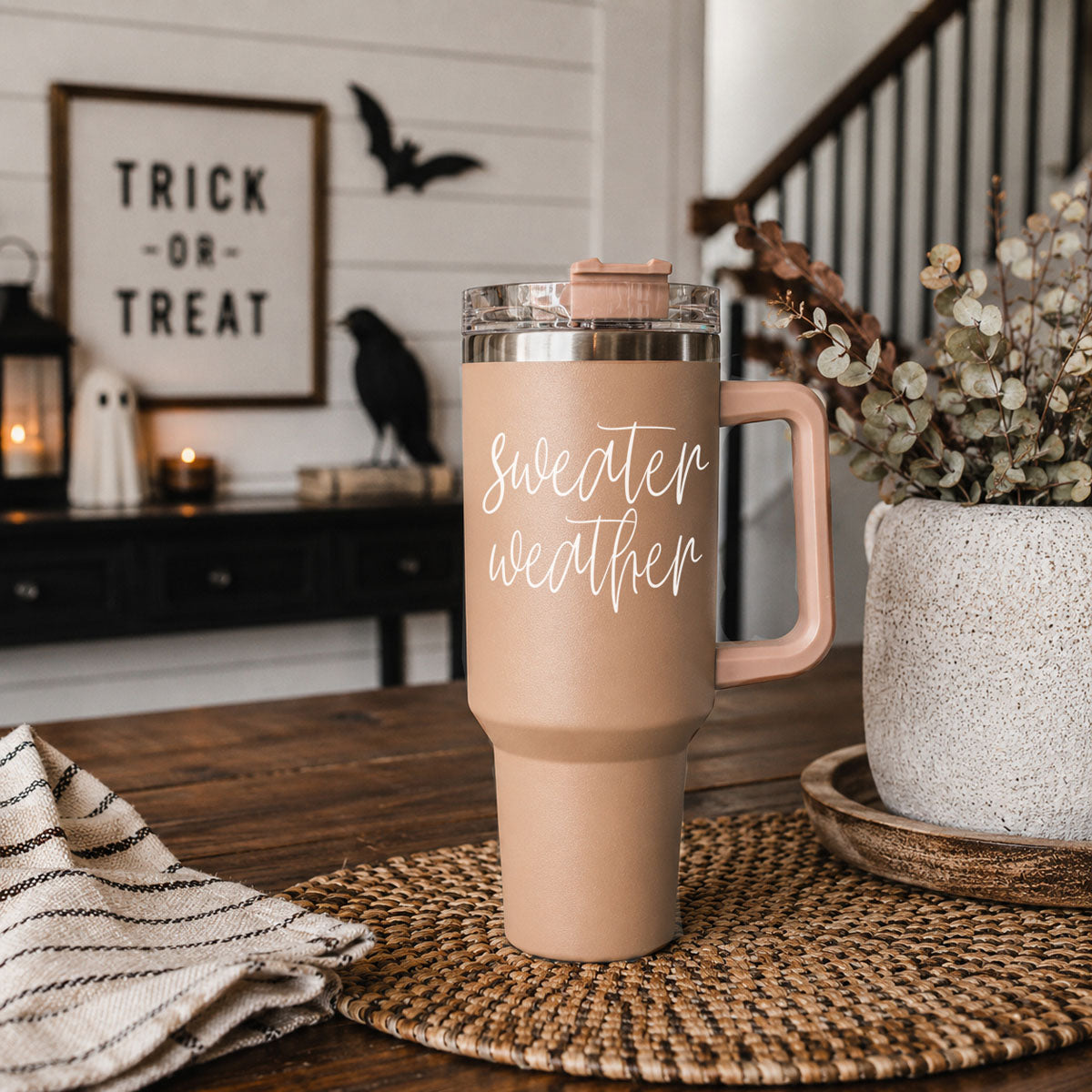Pink travel mug with 'Sweater weather' text on a wooden table with Halloween decorations in the background.