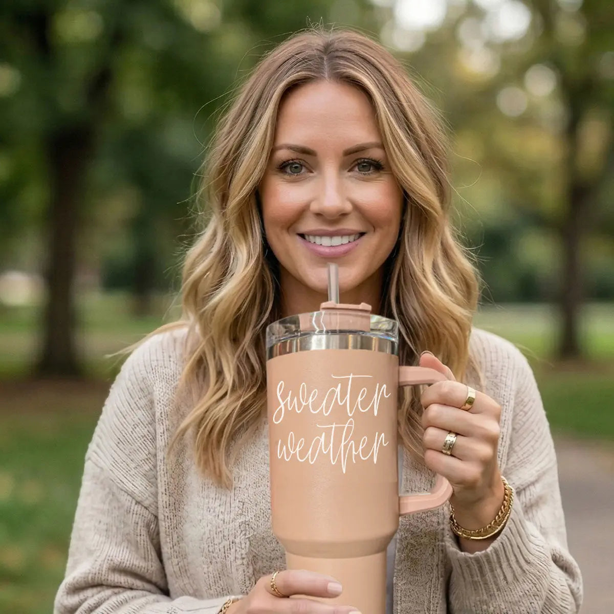 Woman holding a pink insulated tumbler with 'sweater weather' text in a park setting