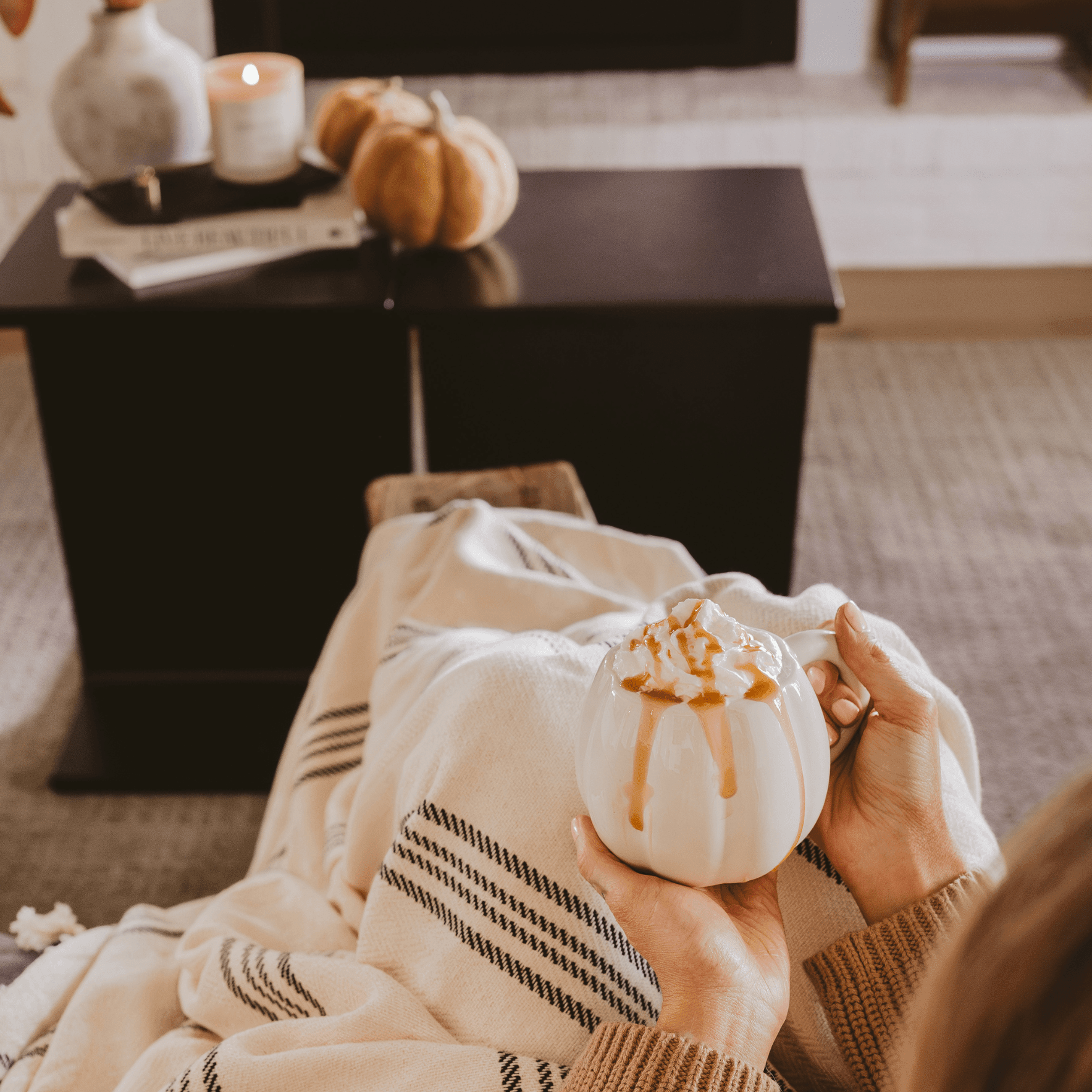 Person holding a small white pumpkin with brown drizzle, sitting on a couch in a cozy living room.
