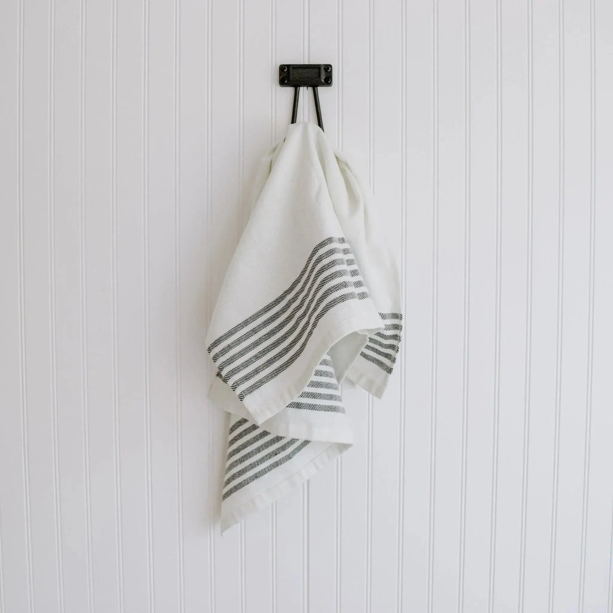 White farmhouse kitchen towel with black stripes hanging on black hook against white beadboard wall