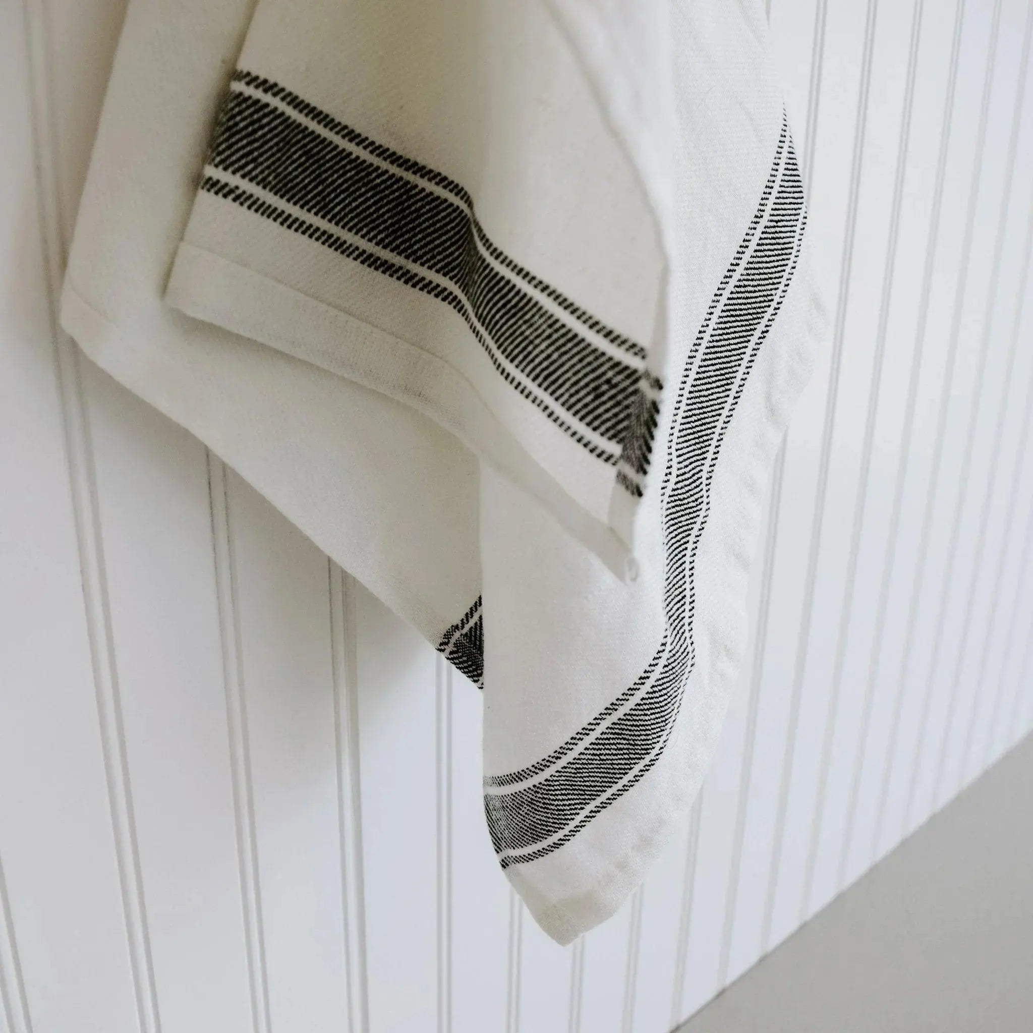 Cream farmhouse kitchen towel with black stripe pattern hanging on white beadboard wall