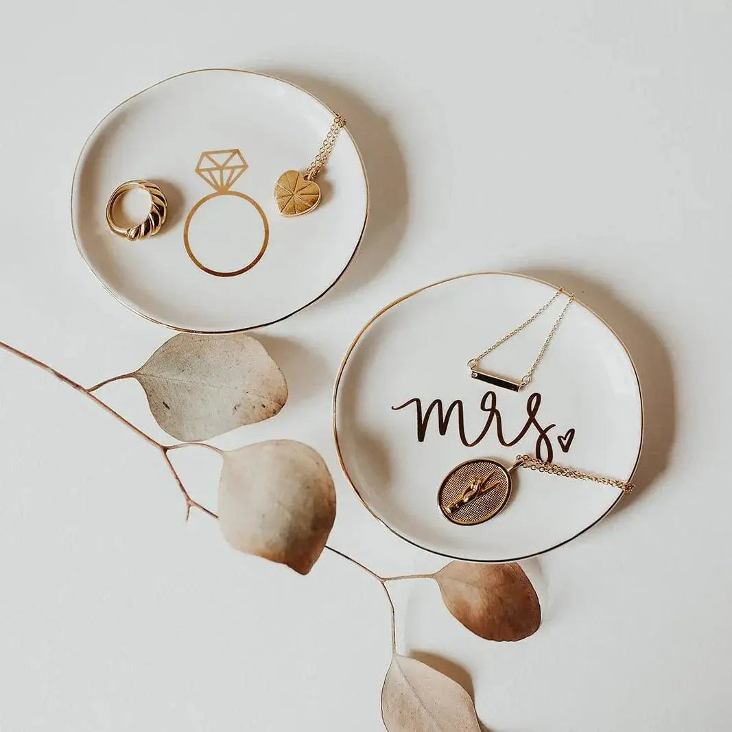 Gold-accented ceramic dishes with 'Mrs' and ring designs, holding delicate necklaces and rings, styled with dried leaves for cozy home decor.