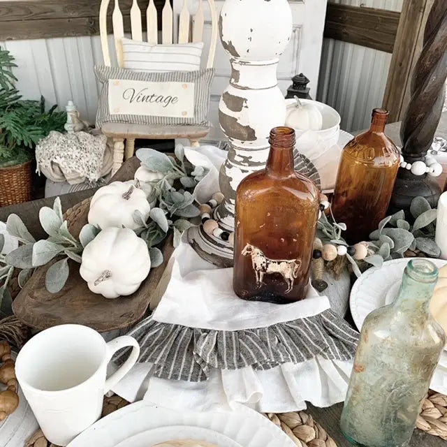 A styled rustic farmhouse table setting featuring amber glass bottles, white ceramic mugs and pumpkins, green eucalyptus leaves, and a striped ruffled tablecloth. A small wooden chair in the background has a pillow that reads 'Vintage'.