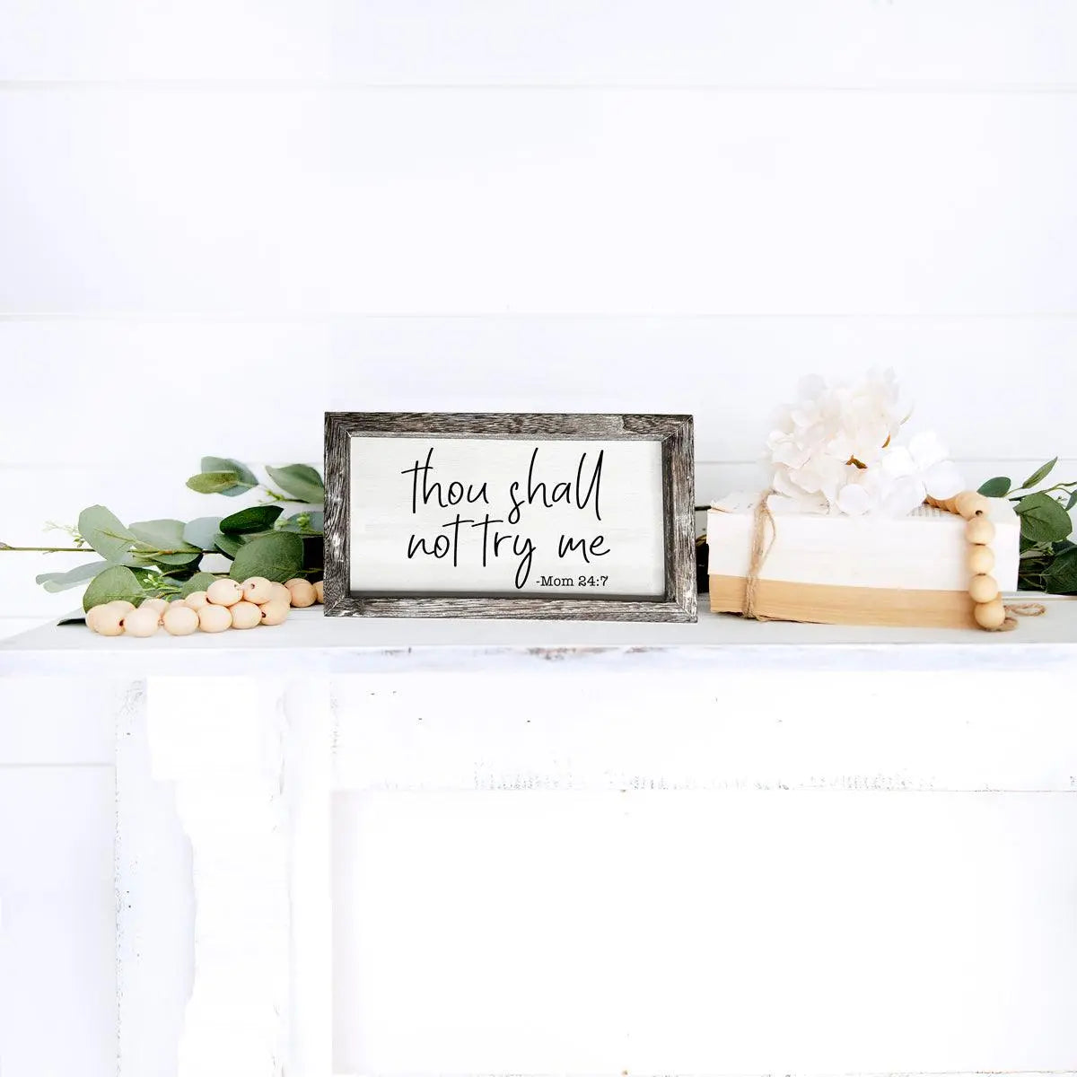 Decorative sign with 'thou shall not try me' text on a white surface with greenery and candles.