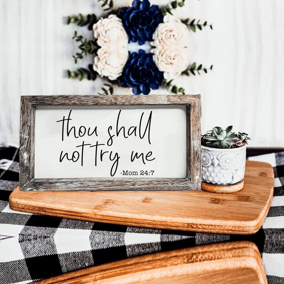 Decorative sign with 'Thou shall not try me' text on a wooden surface with flowers in the background.