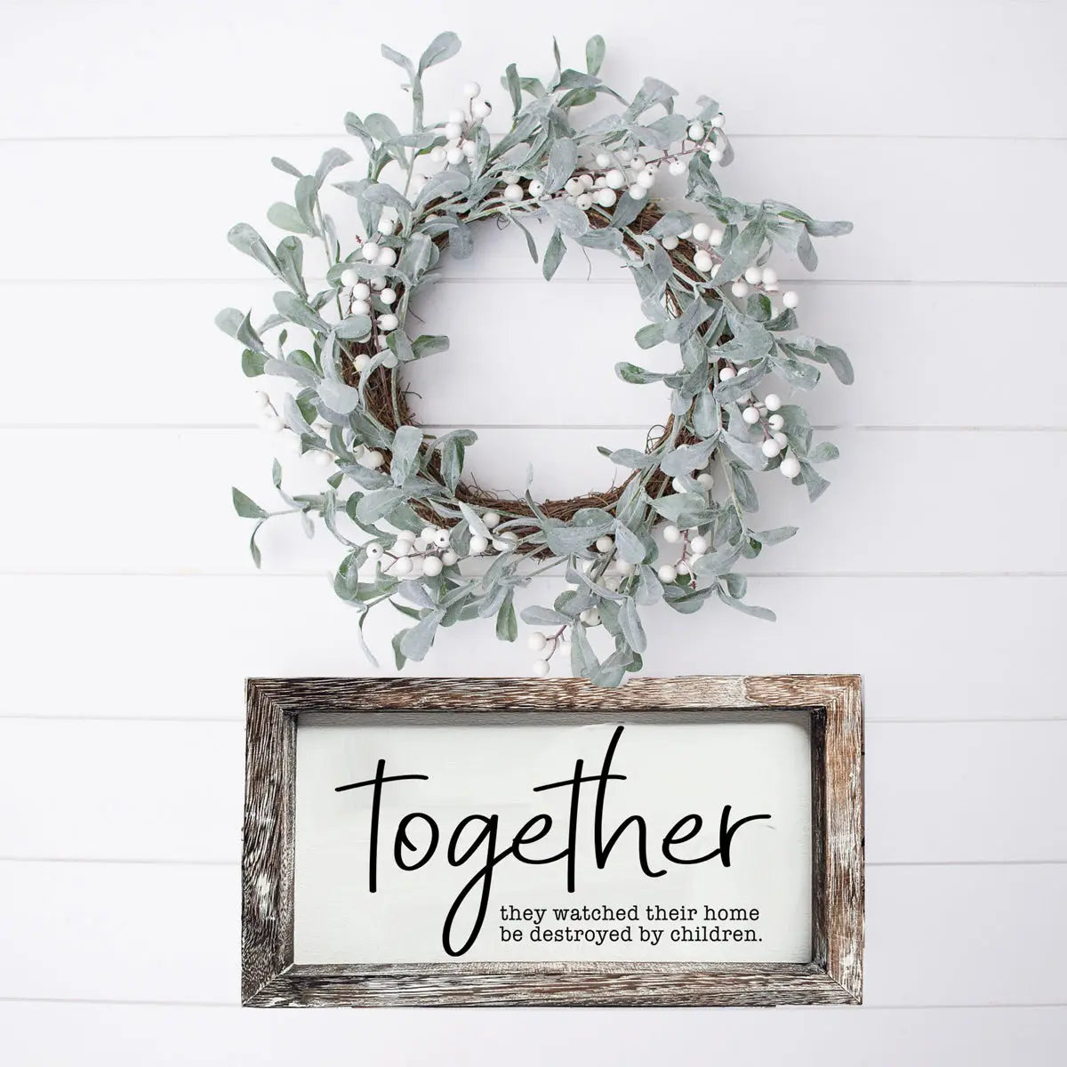 Decorative wreath with greenery and a framed sign on a white wooden background