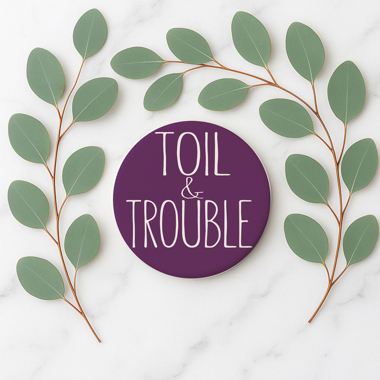 Purple circular sign with 'Toil & Trouble' text surrounded by green leaves on a white marble background