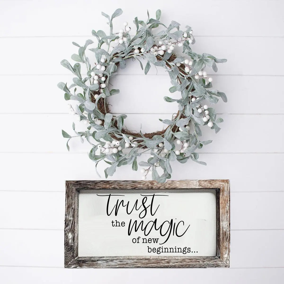 Wreath with white berries on a white wooden wall above a framed sign with text.