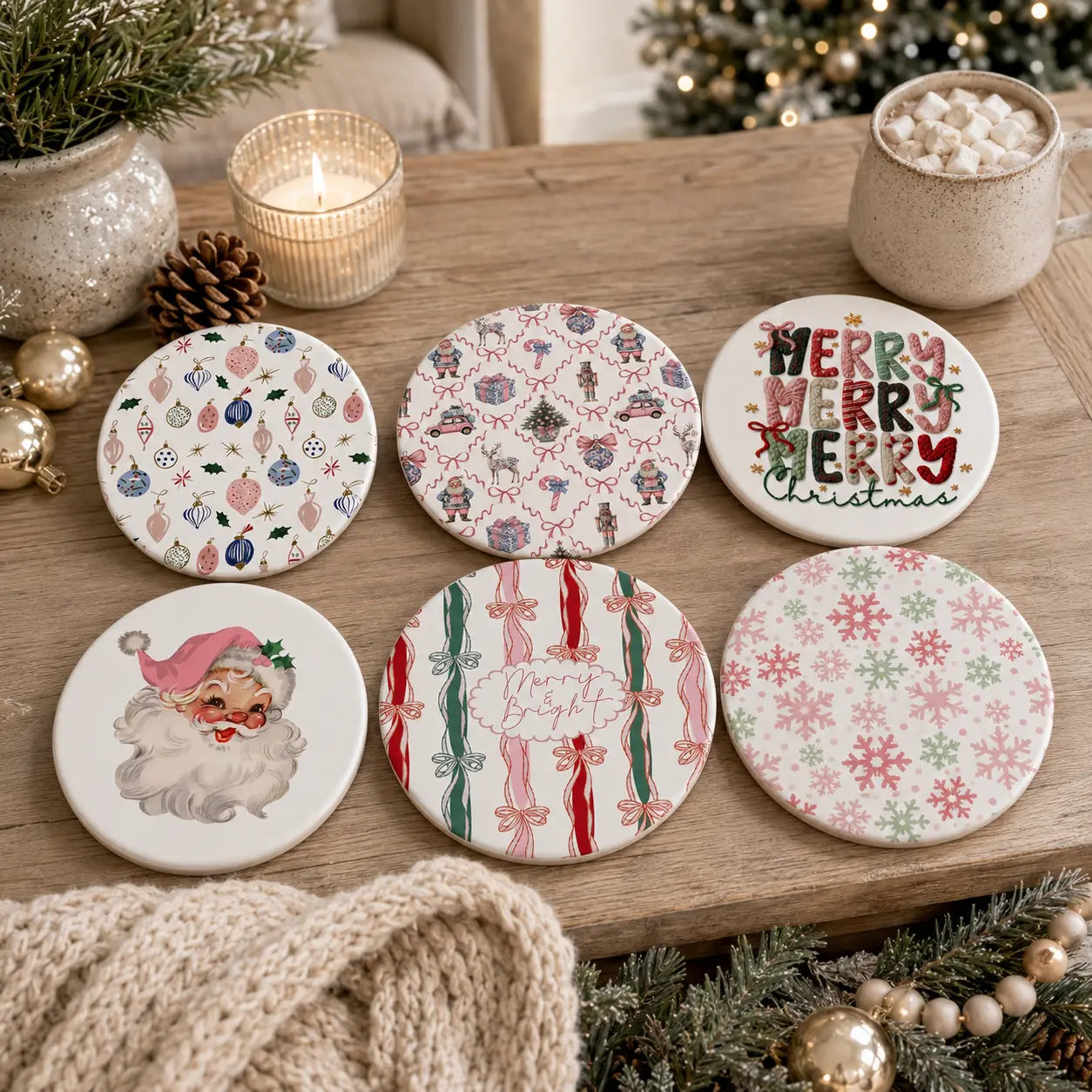 Six round ceramic coasters with festive Christmas designs are arranged on a wooden table surrounded by holiday decorations, including a candle, pinecones, a pot of marshmallows, and evergreen branches.