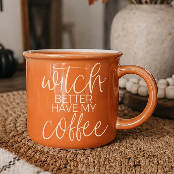 An orange ceramic coffee mug with the phrase 'witch BETTER HAVE MY coffee' in white script and block lettering, resting on a woven brown placemat with a blurred home interior background.