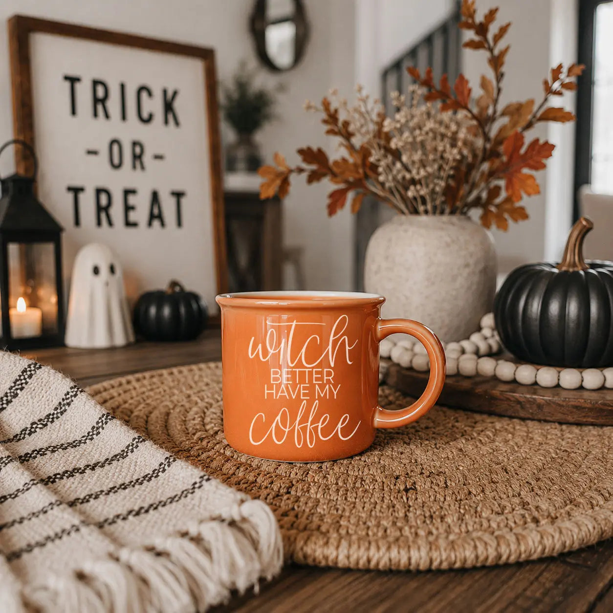 An orange ceramic mug with white text reading 'I RUN ON pumpkin spice, HORROR MOVIES cuss words' sits on a woven placemat in a cozy autumn-themed setting with pumpkins, a sign reading 'Trick or Treat', and a candle.