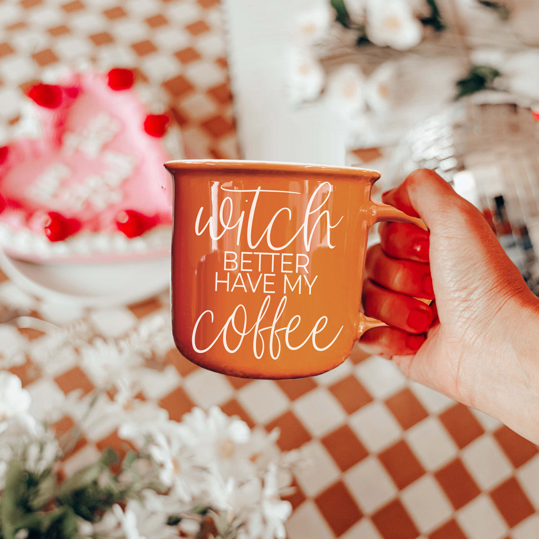 Hand holding an orange mug with 'Witch better have my coffee' text against a checkered tablecloth background.
