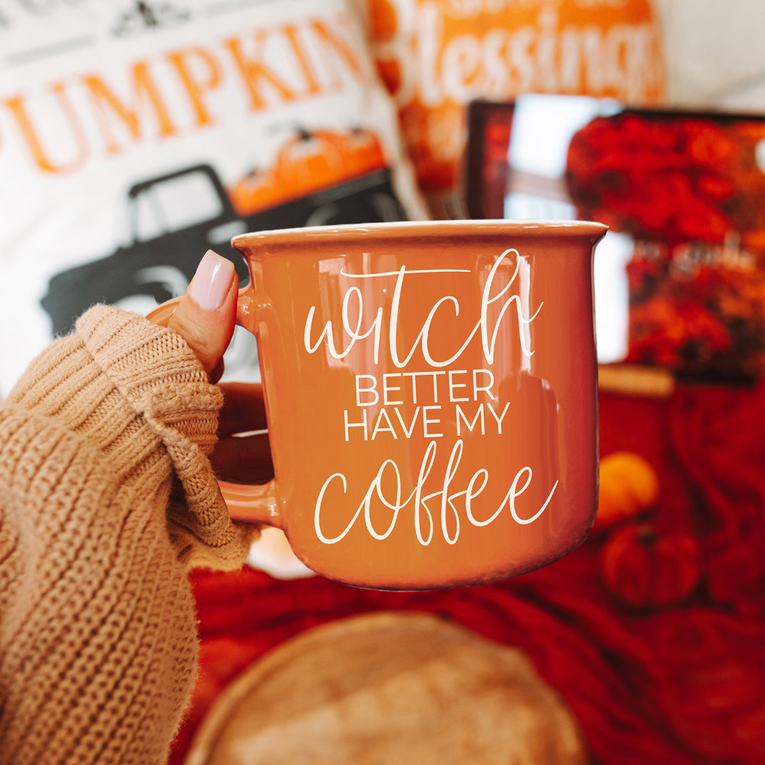 Person holding an orange mug with text, surrounded by autumn-themed decor
